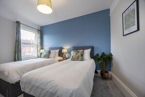 A hotel room with two single beds, a large window with curtains, a potted plant, and a framed picture on the wall.