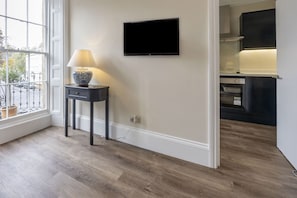 A room with a TV mounted on the wall, a small table with a lamp, and a view through a window to the outside. - Elegant Period Town Centre Apartment with Parking (Cheltenham)