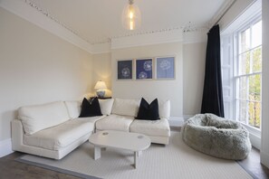 Ett modernt vardagsrum med en vit soffa, ett vitt soffbord och en mjuk sittpuff. På väggen hänger tre inramade tavlor, och på ett sidobord står en lampa.