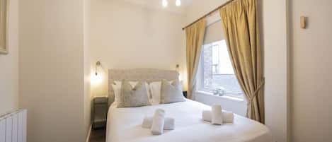 A neatly made bed with white linens and pillows, a modern chandelier, and a window with beige curtains.