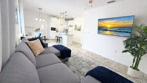 Een moderne woonkamer met een grijze hoekbank, een eethoek met een keuken op de achtergrond, en een groot schilderij van een zonsondergang aan de muur.