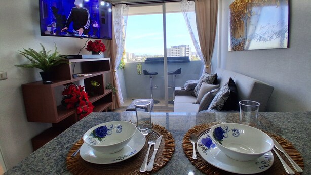 Een moderne woonkamer met een flatscreen televisie, een bank, een eethoek met twee kommen en bestek, en een balkon met uitzicht.