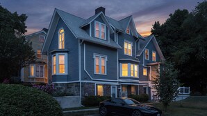 Une grande maison à plusieurs étages, dotée d’un toit à pignon bien visible, de nombreuses fenêtres et d’une allée où une voiture est garée.