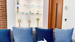 A living room with a grey sofa, blue patterned cushions, a glass shelf with decorative items, and white orchids on a tiled floor. - La Dimora Holiday House (Salerno)