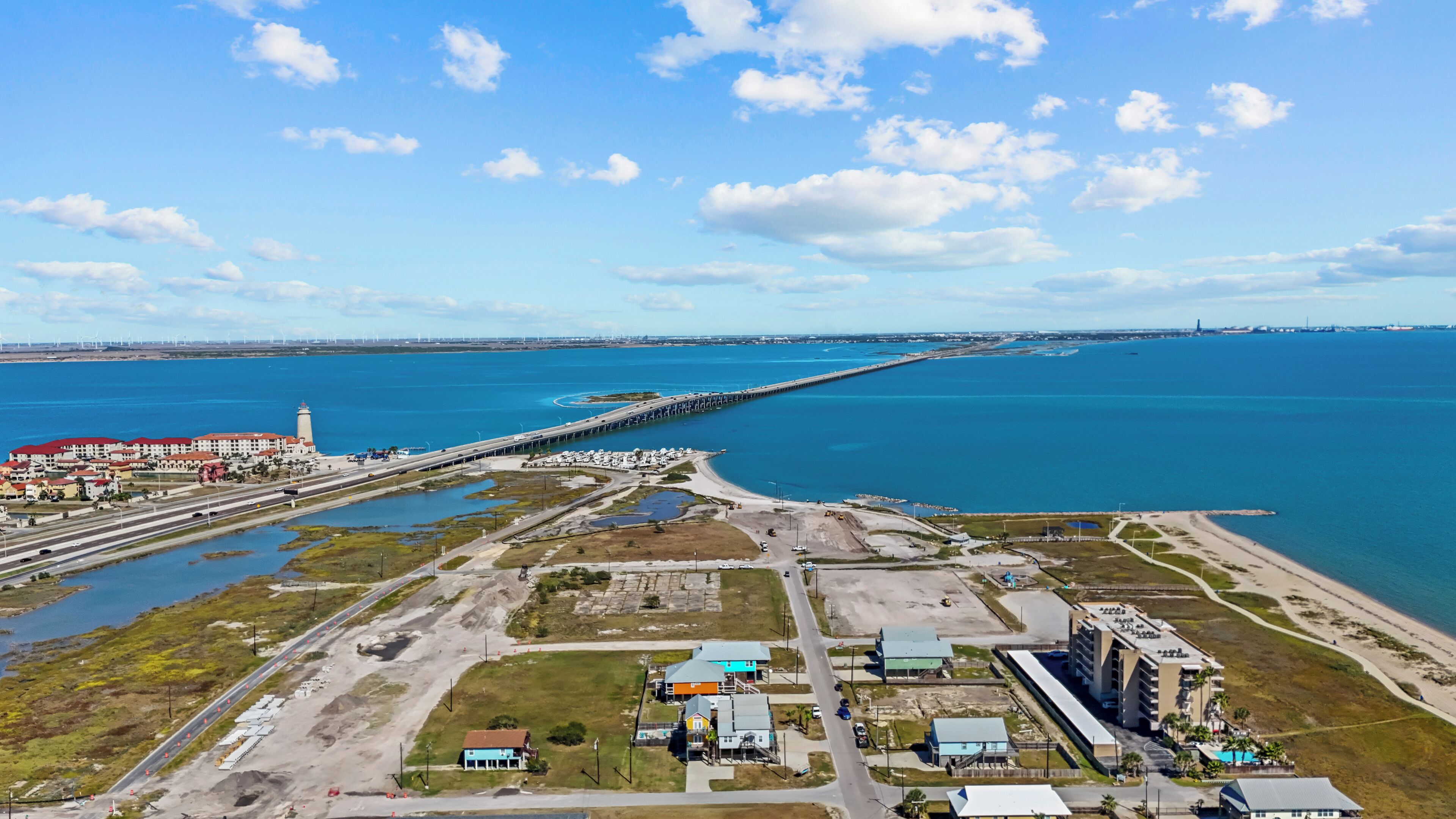 An aerial view of a coastal area with a long pier extending into the water, a road leading to a building complex, and a beach in the background.