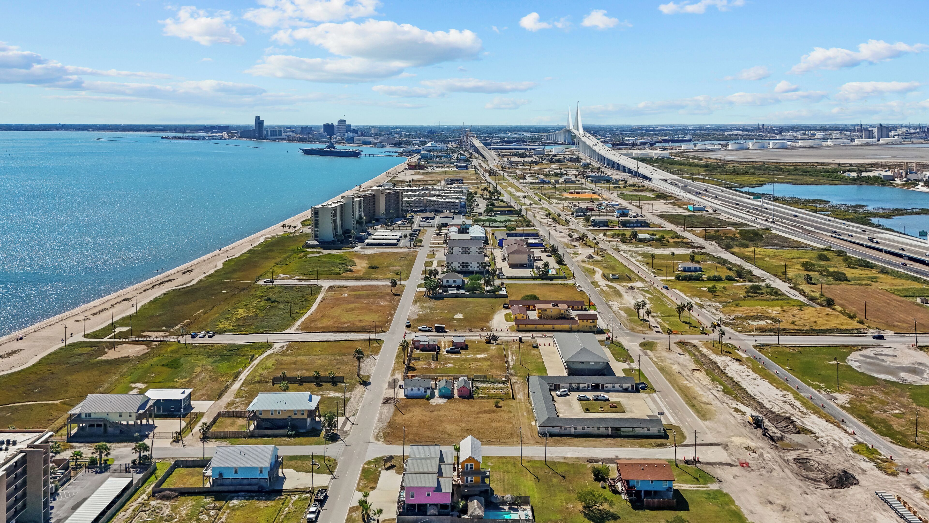 Vista aérea de una zona costera con un puente grande, un puerto deportivo y varios edificios, incluyendo un hotel con piscina y un estacionamiento.