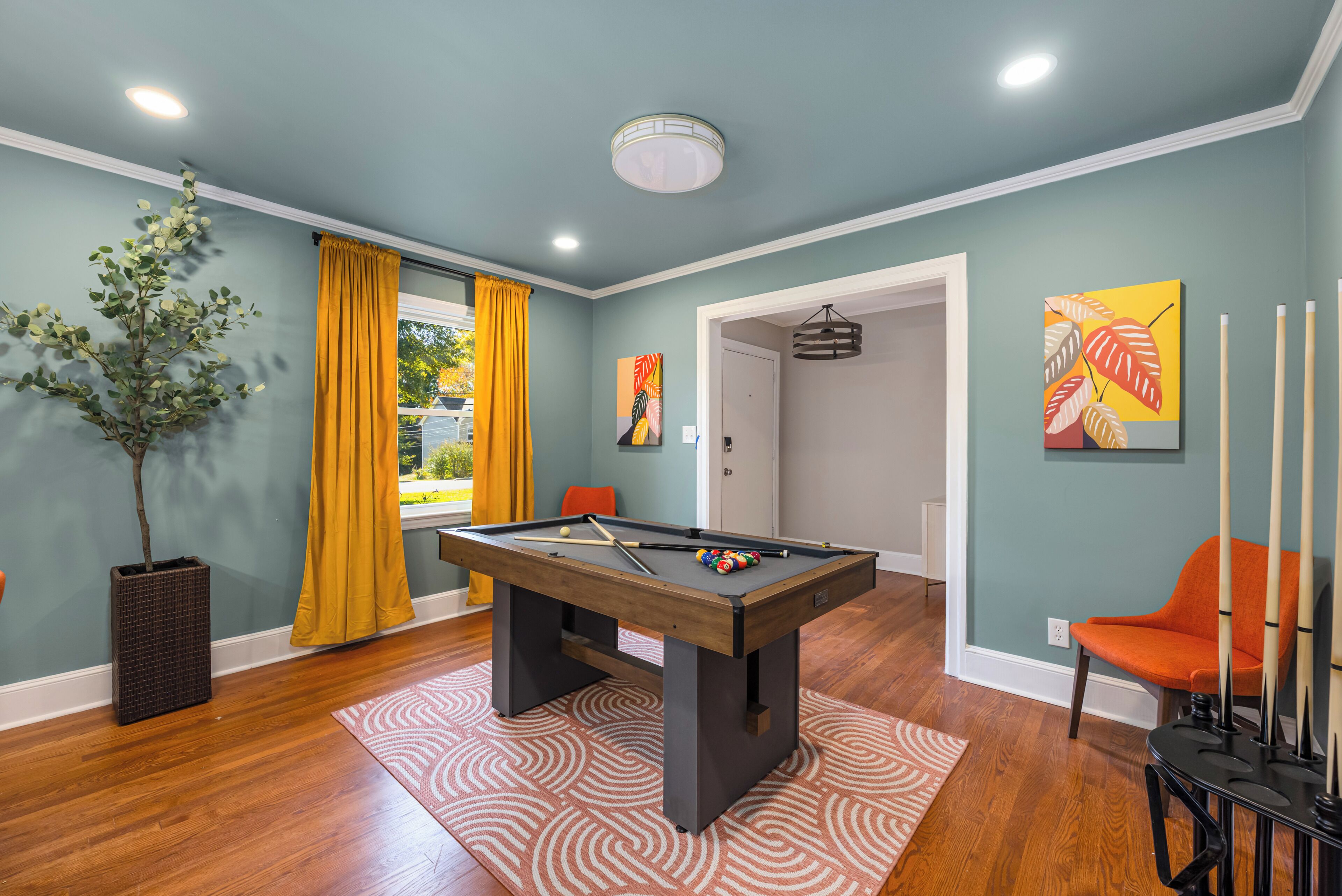A pool table in a room with teal walls, a potted plant, and a yellow curtain.