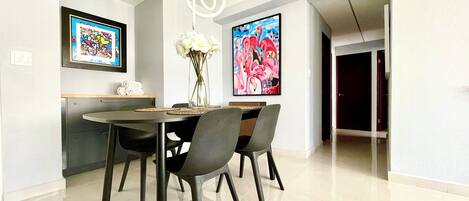 A modern dining area with a round table, black chairs, and a vase of flowers. There are two framed artworks on the walls and a ceiling light fixture.