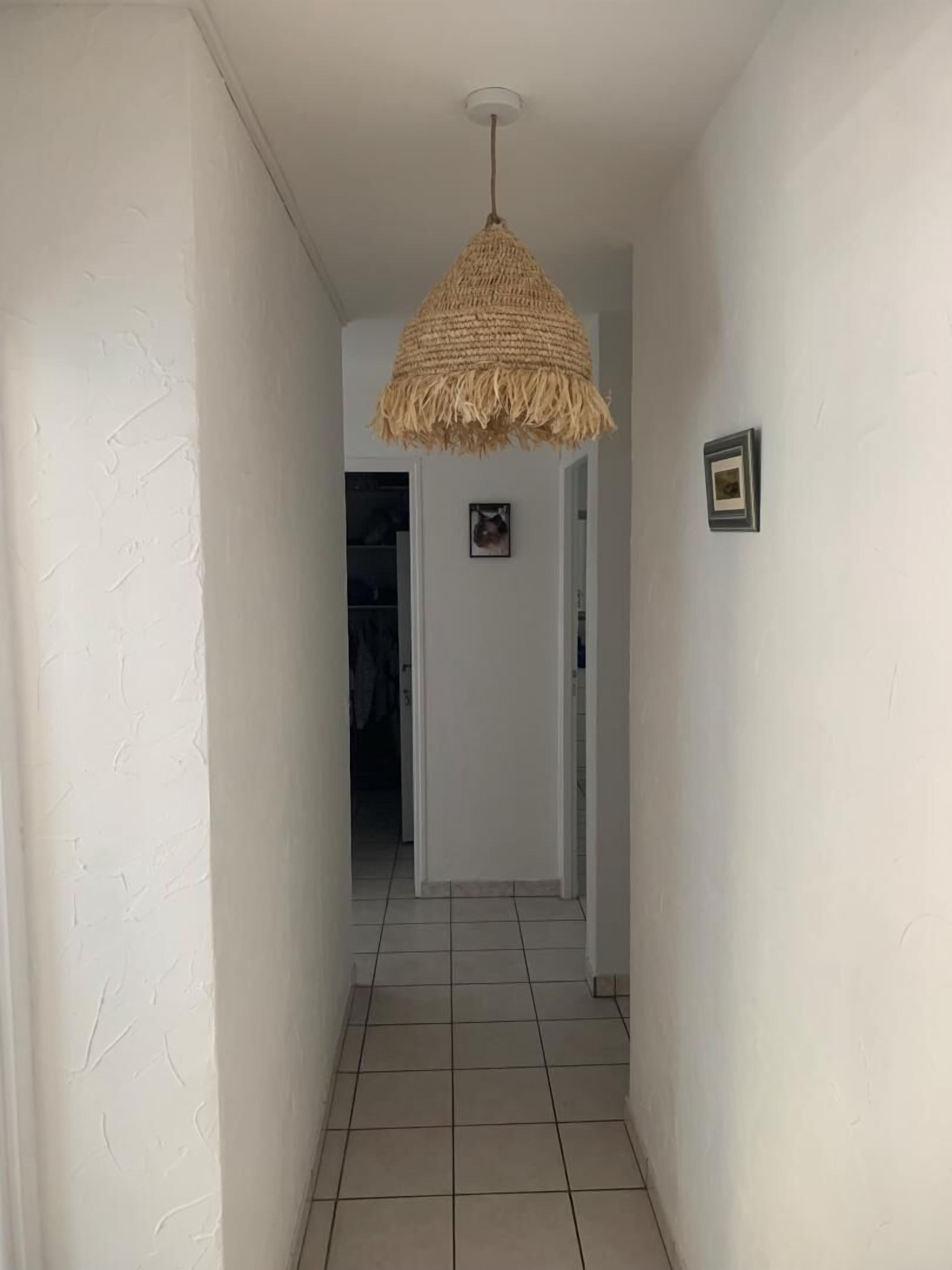 A hallway with white walls and a tiled floor, leading to a room with a door. A woven pendant light hangs from the ceiling.