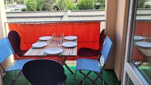 En balkong med ett träbord dukat för fyra personer, omgiven av blå stolar, och en röd gardin i bakgrunden.