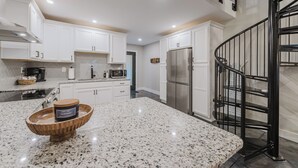 A modern kitchen with white cabinets, granite countertops, a stainless steel refrigerator, and a black spiral staircase.