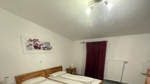 A bedroom with two wooden beds, a nightstand with a lamp, a radiator, and a window with a red curtain. - Attika Hotel (Halstenbek)