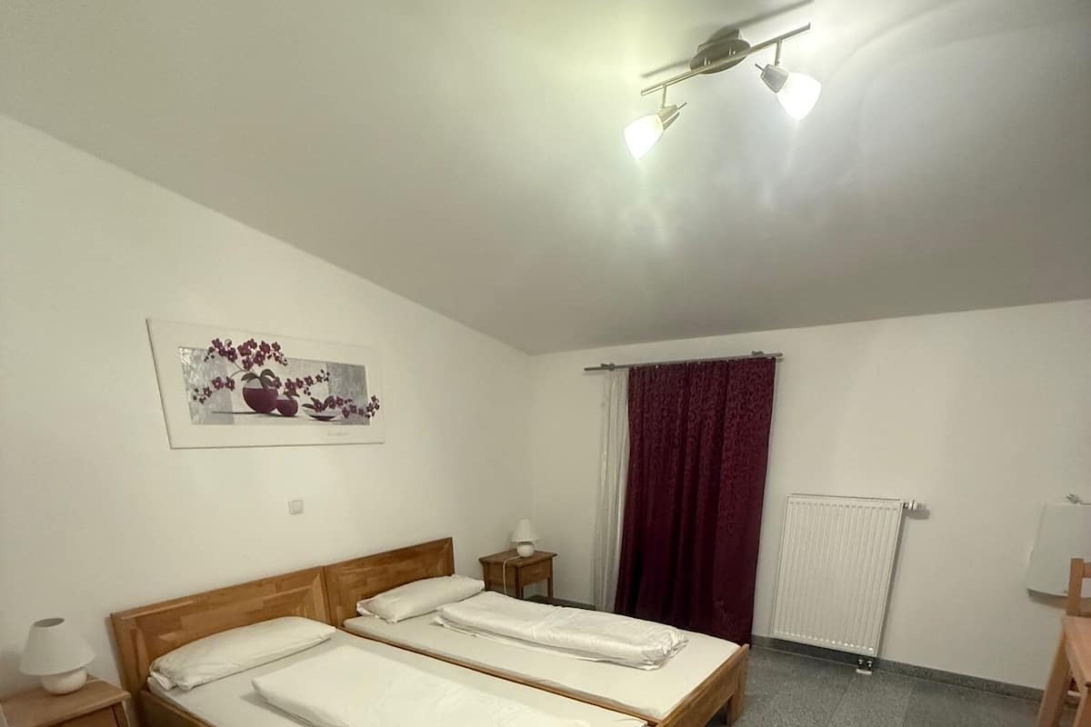 Un dormitorio con dos camas de madera, una mesita de noche con lámpara, un radiador y una ventana con cortina roja.