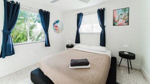 Una camera da letto con un letto, una ventola a soffitto, una finestra con tende, un comodino e un quadro appeso al muro.