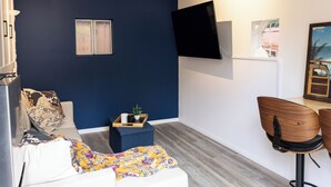 Een kamer met een blauwe wand, een wandgemonteerde televisie, een witte bank met decoratieve kussens, een klein houten bijzettafeltje en een houten stoel.