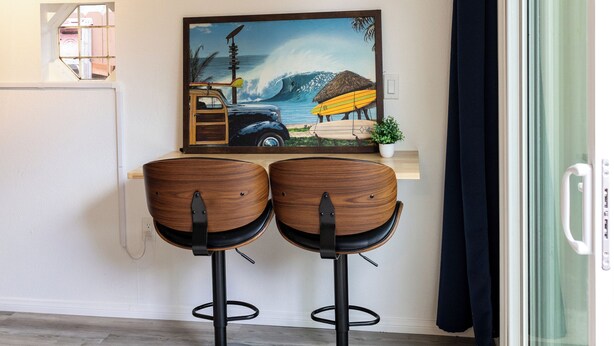Een moderne kamer met een glazen deur, twee houten barstoelen, een schilderij van een strand en een kleine potplant.