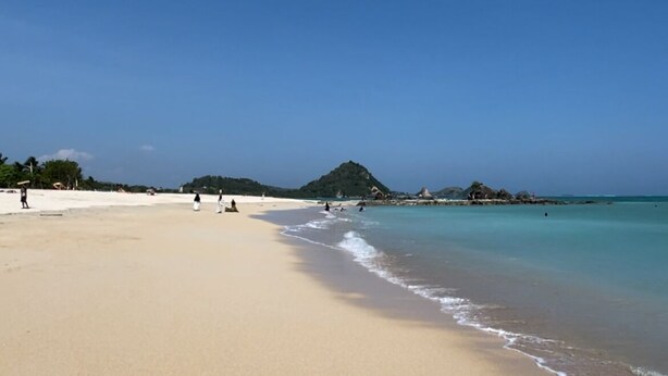 Ein weiter Sandstrand mit sanften Wellen, blauem Himmel und einem fernen Berg.