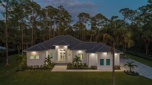 A modern two-story house with a well-maintained lawn, a driveway, and a large front yard with palm trees. - The Naples Luxe Retreat . Game Room • Heated Pool (Naples)