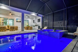 Et moderne poolområde indendørs med en blålig pool, et loungeområde med træmøbler og et køkken i baggrunden.
