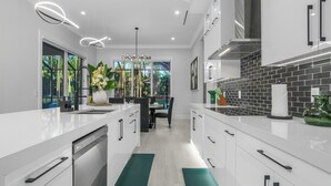 A modern kitchen with white cabinetry, a black tiled backsplash, and a dining area with a view of trees. - The Naples Luxe Retreat . Game Room • Heated Pool (Naples)