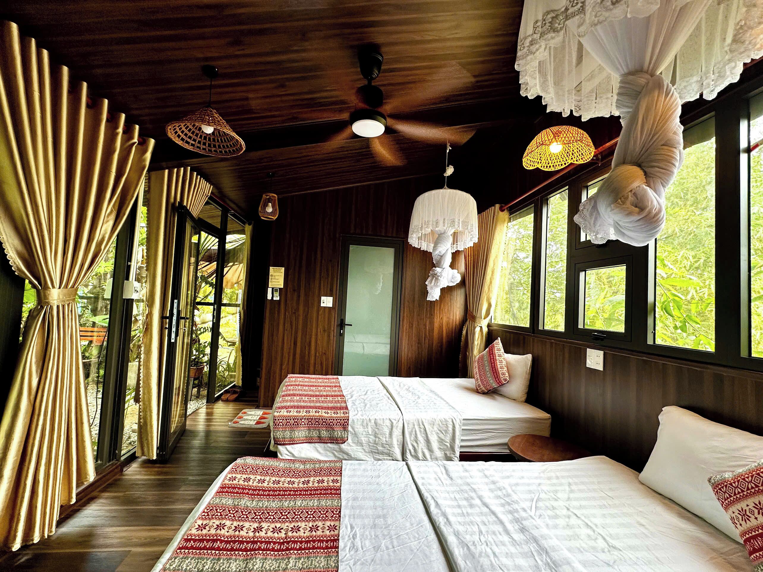Una habitación con paredes de madera, ventilador de techo, dos camas con ropa de cama estampada y ventanas amplias con cortinas.