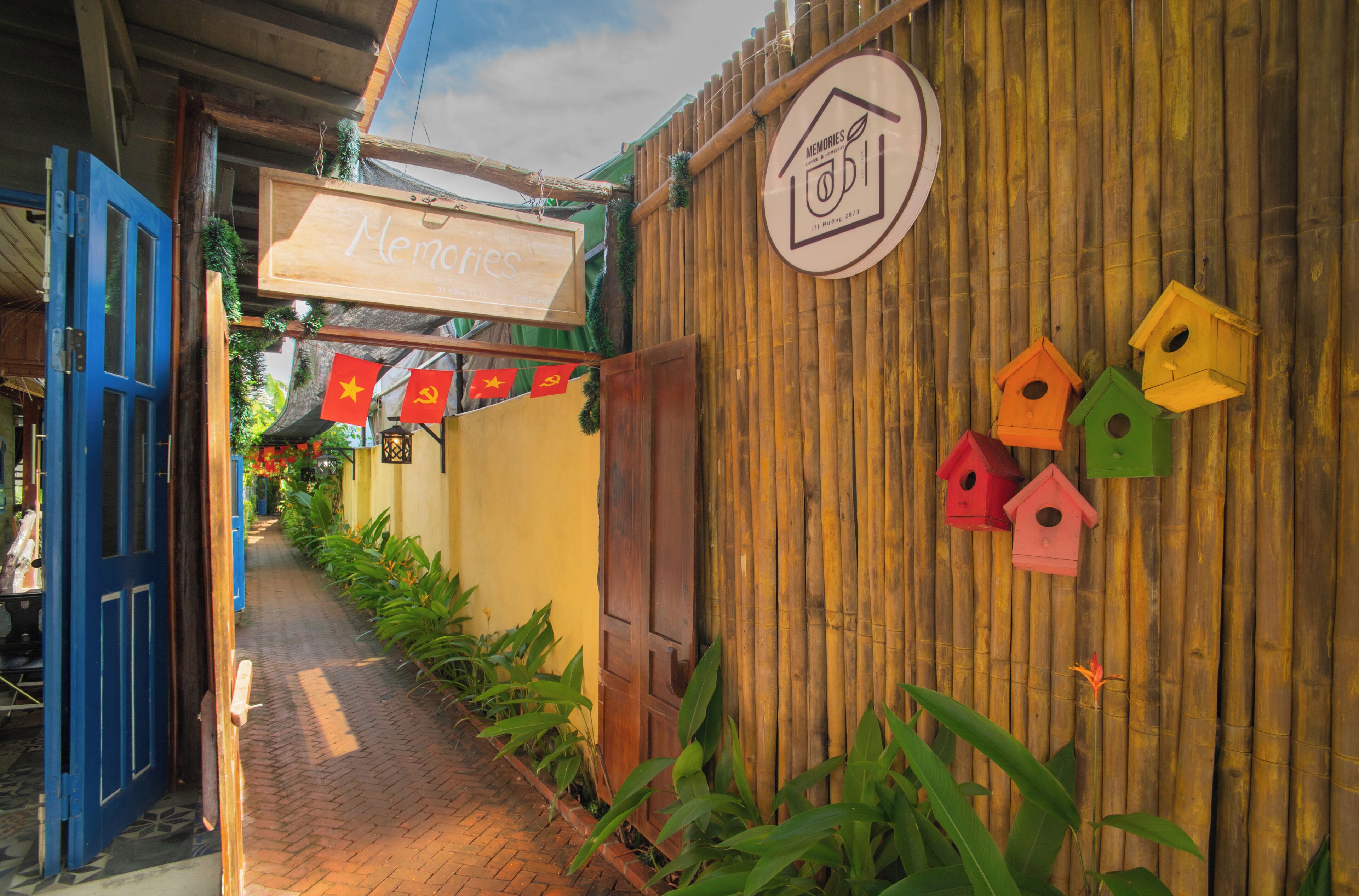 Une ruelle étroite avec une porte en bois, une enseigne indiquant « Souvenirs », et des nichoirs colorés accrochés à un mur en bambou.