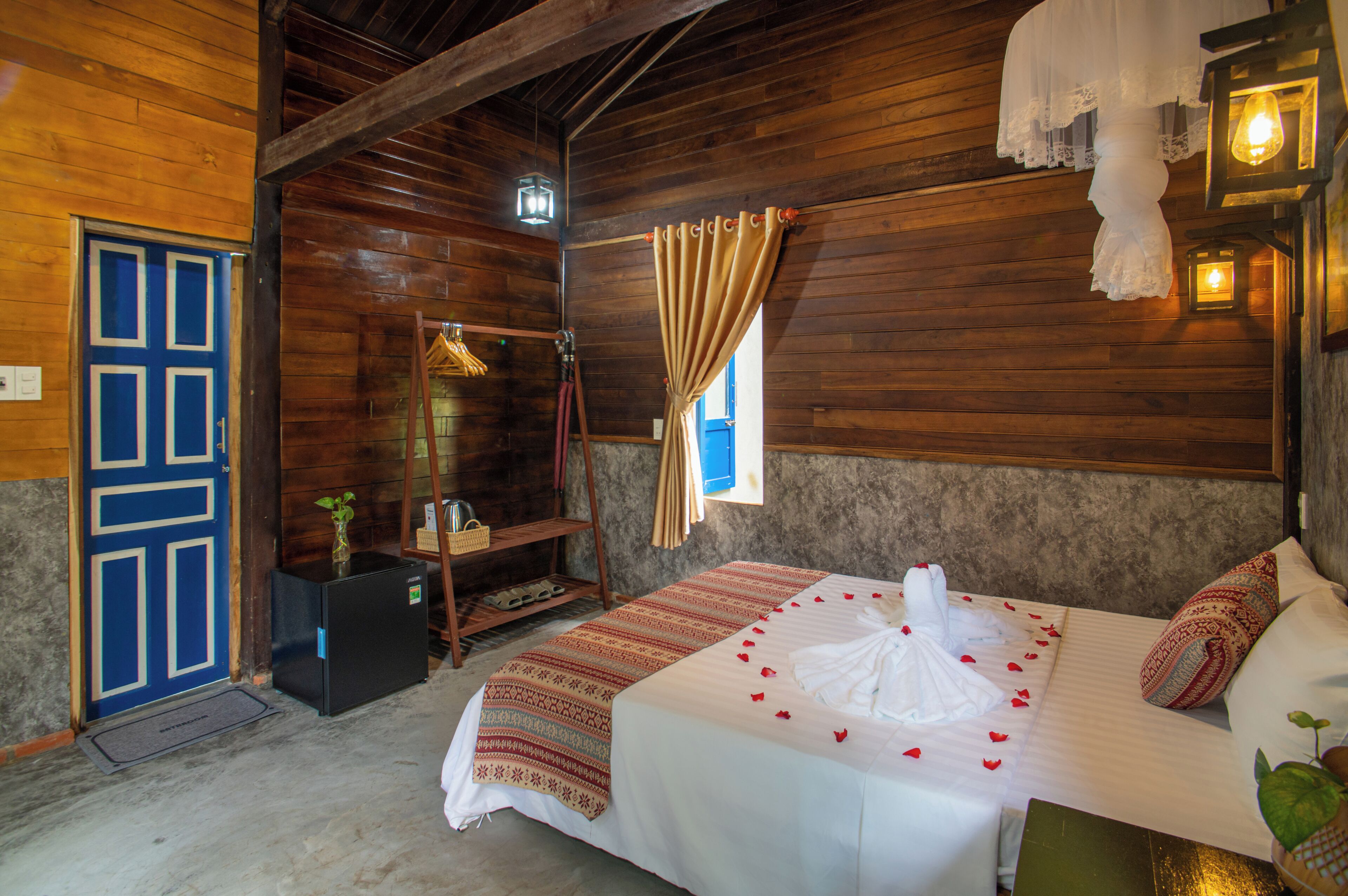 A wooden room with a bed, a small table, a door, and a window with curtains.