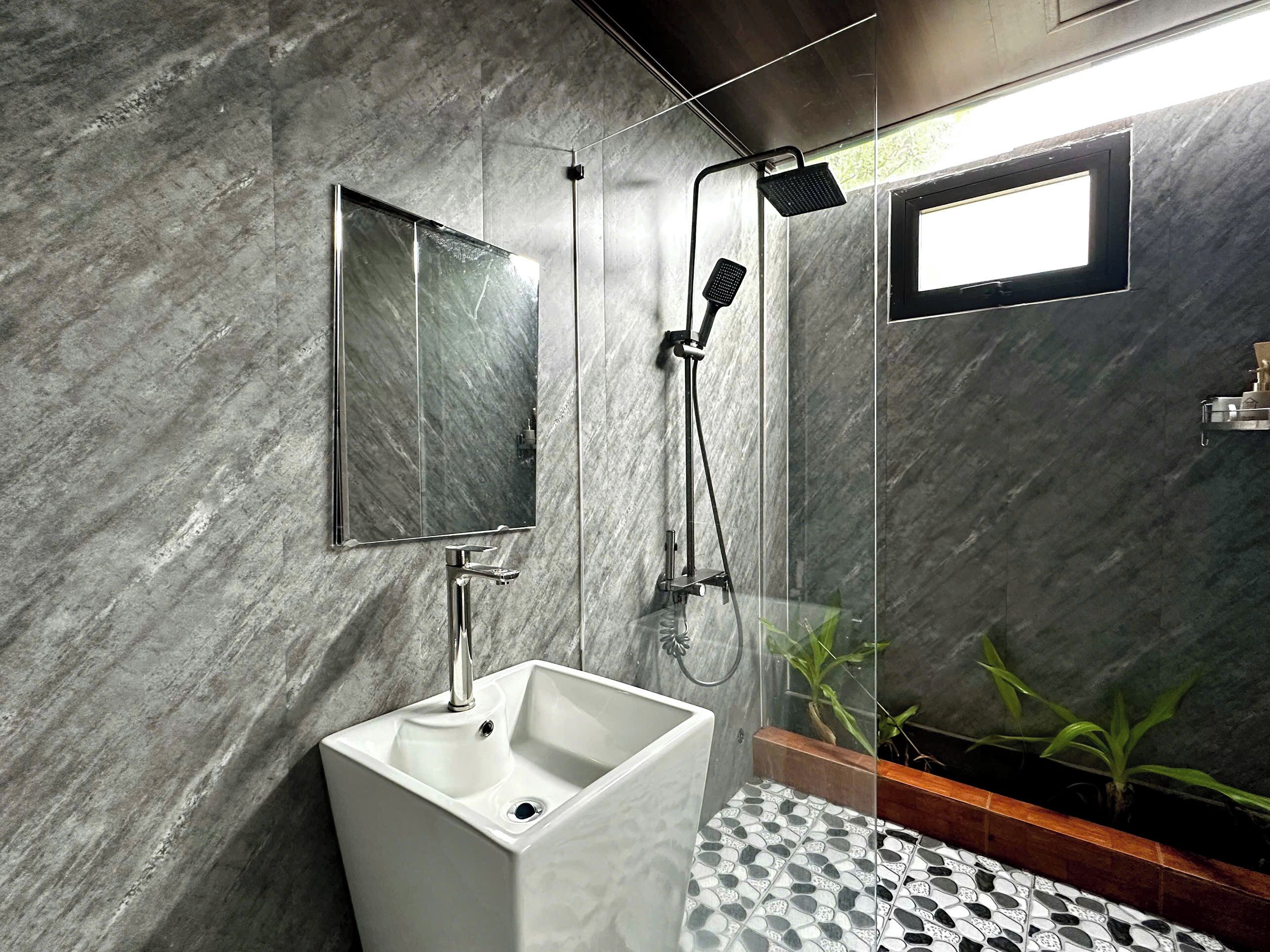 A modern bathroom with a marble wall, a white sink, a mirror, a shower, and a small window.