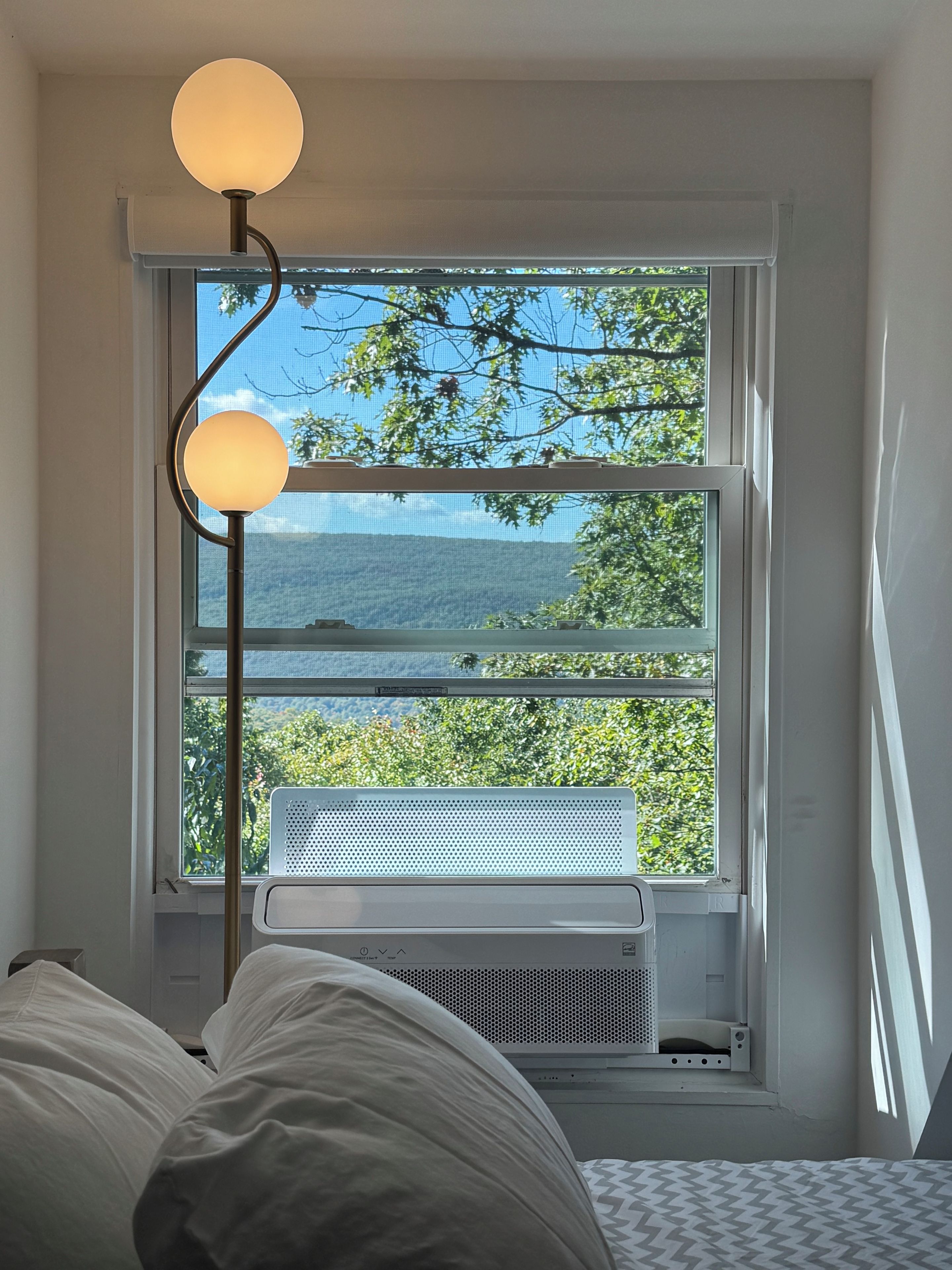 A bedroom with a bed, a standing floor lamp, a window with a view of greenery and a body of water.