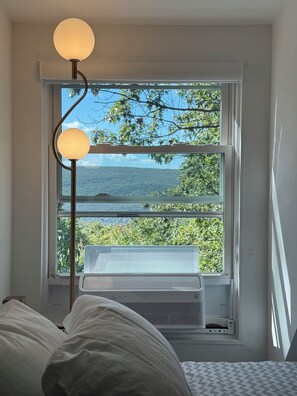Un dormitorio con cama, una lámpara de pie, una ventana con vistas a follaje y una masa de agua.