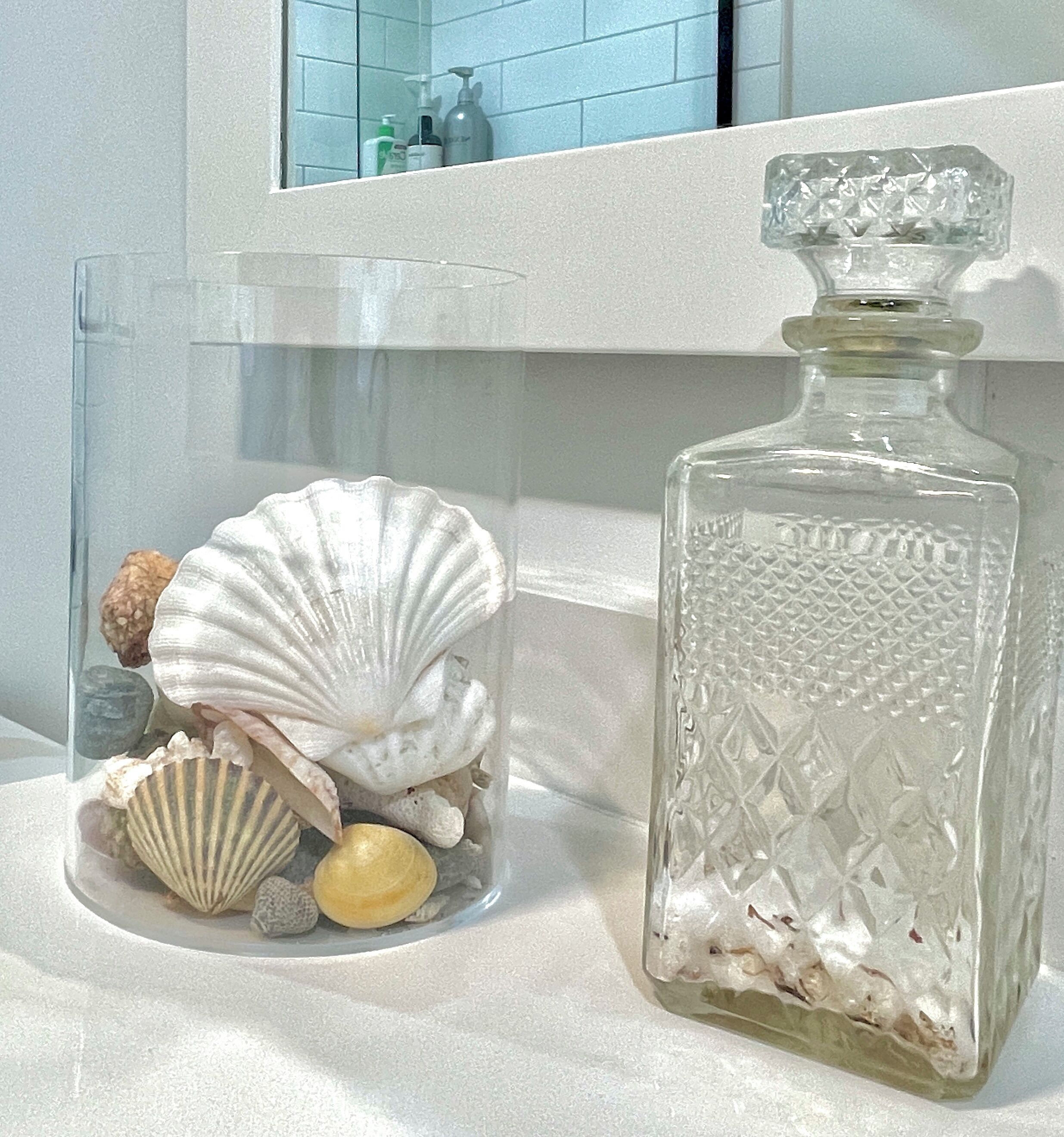 Una botella de vidrio transparente con diseños en relieve, un frasco de vidrio con conchas marinas y una encimera de baño con un espejo y artículos de aseo.