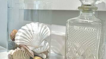 Una botella de vidrio transparente con diseños en relieve, un frasco de vidrio con conchas marinas y una encimera de baño con un espejo y artículos de aseo.