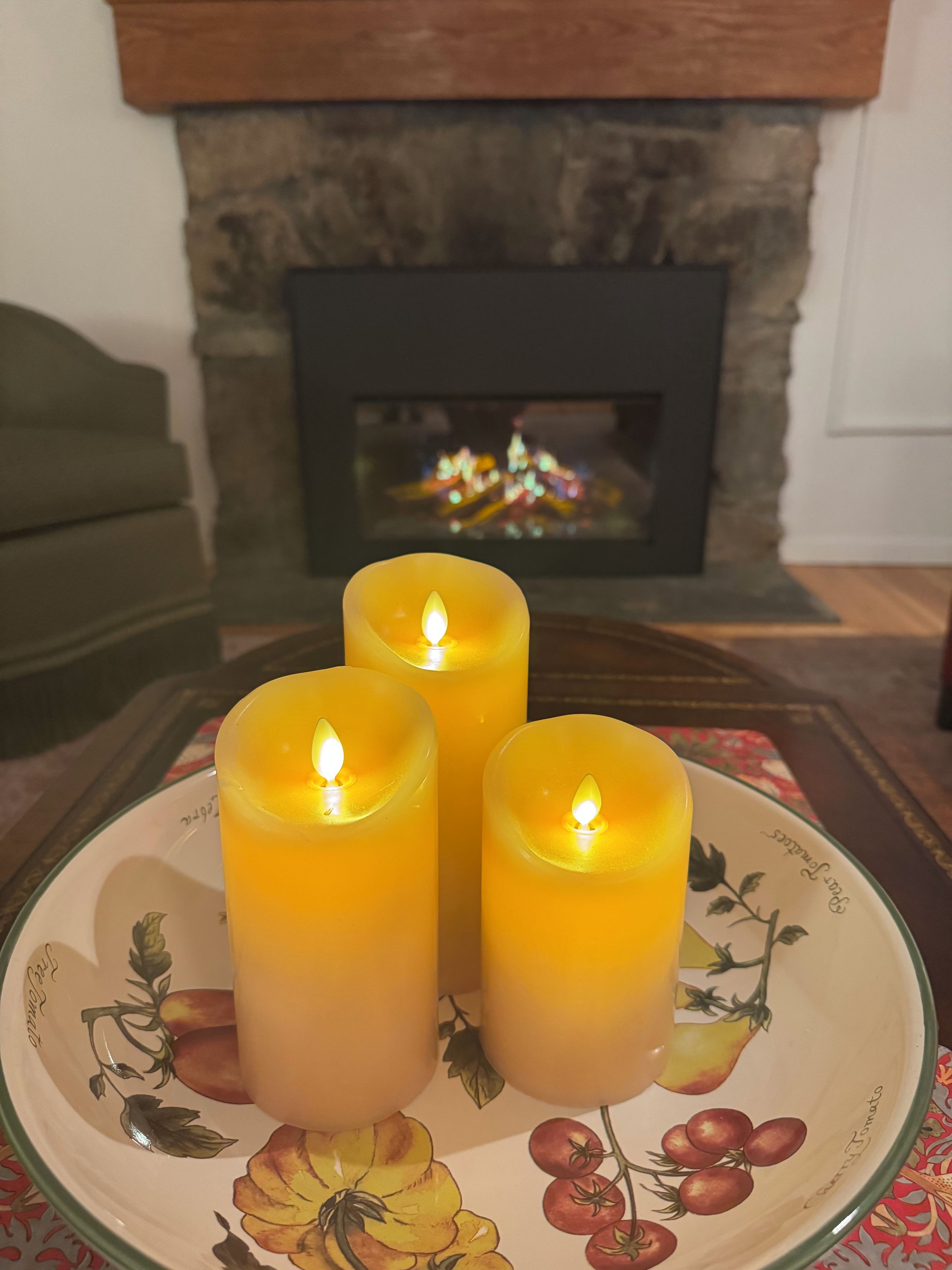 Three candles on a plate with fruit designs, a fireplace in the background, and a green sofa.
