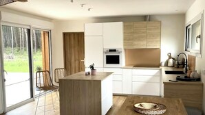 Een moderne keuken met een centraal eiland, witte kasten en uitzicht op een bos door grote schuifdeuren.