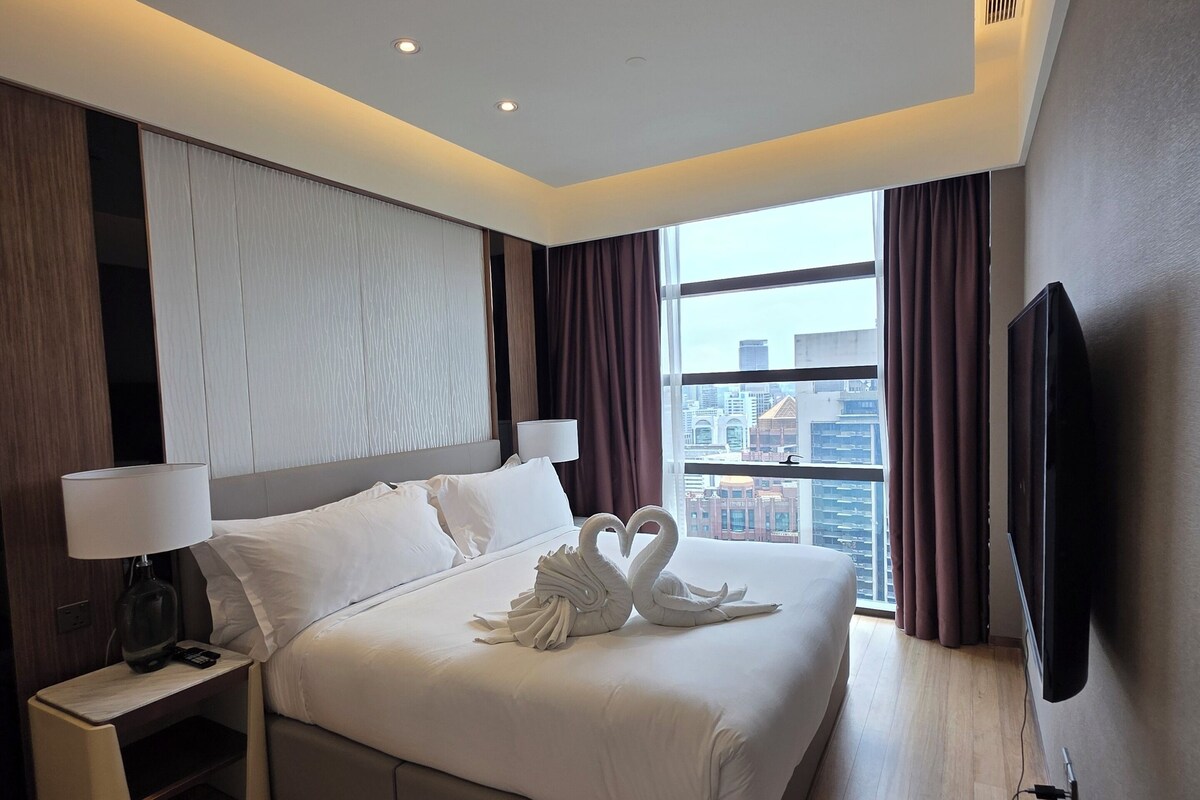 Une chambre d’hôtel moderne dotée d’un grand lit, d’un téléviseur à écran plat, d’une table de chevet avec une lampe et offrant une vue sur la ville grâce à de grandes fenêtres.