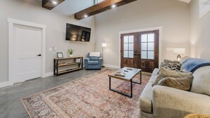 Ein geräumiges Wohnzimmer mit großem Teppich, einer Couch, einem Couchtisch und einem blauen Sessel. Man sieht einen Holzbalken an der Decke, einen an der Wand befestigten Fernseher und eine Tür mit Glasfeldern.