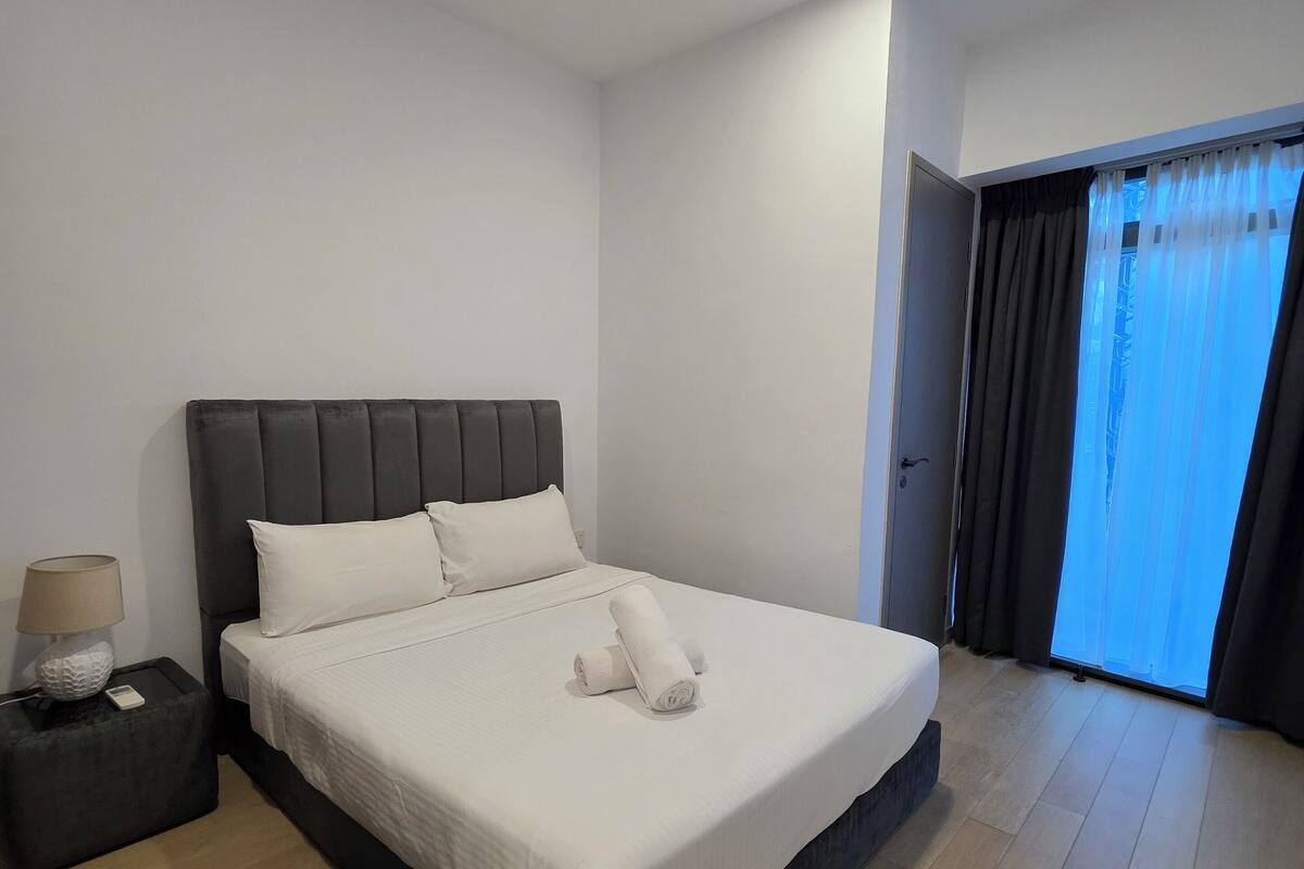 Een moderne slaapkamer met een groot bed, een nachtkastje met een lamp, en uitzicht naar buiten door een raam met gordijnen.