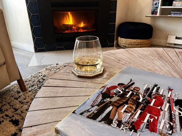 Un magazine posé sur une table en bois, à côté d’un verre de vin blanc, avec une cheminée et une bibliothèque en arrière-plan.