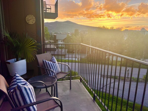 A balcony with chairs and a potted plant, overlooking a road and buildings with a sunset view.