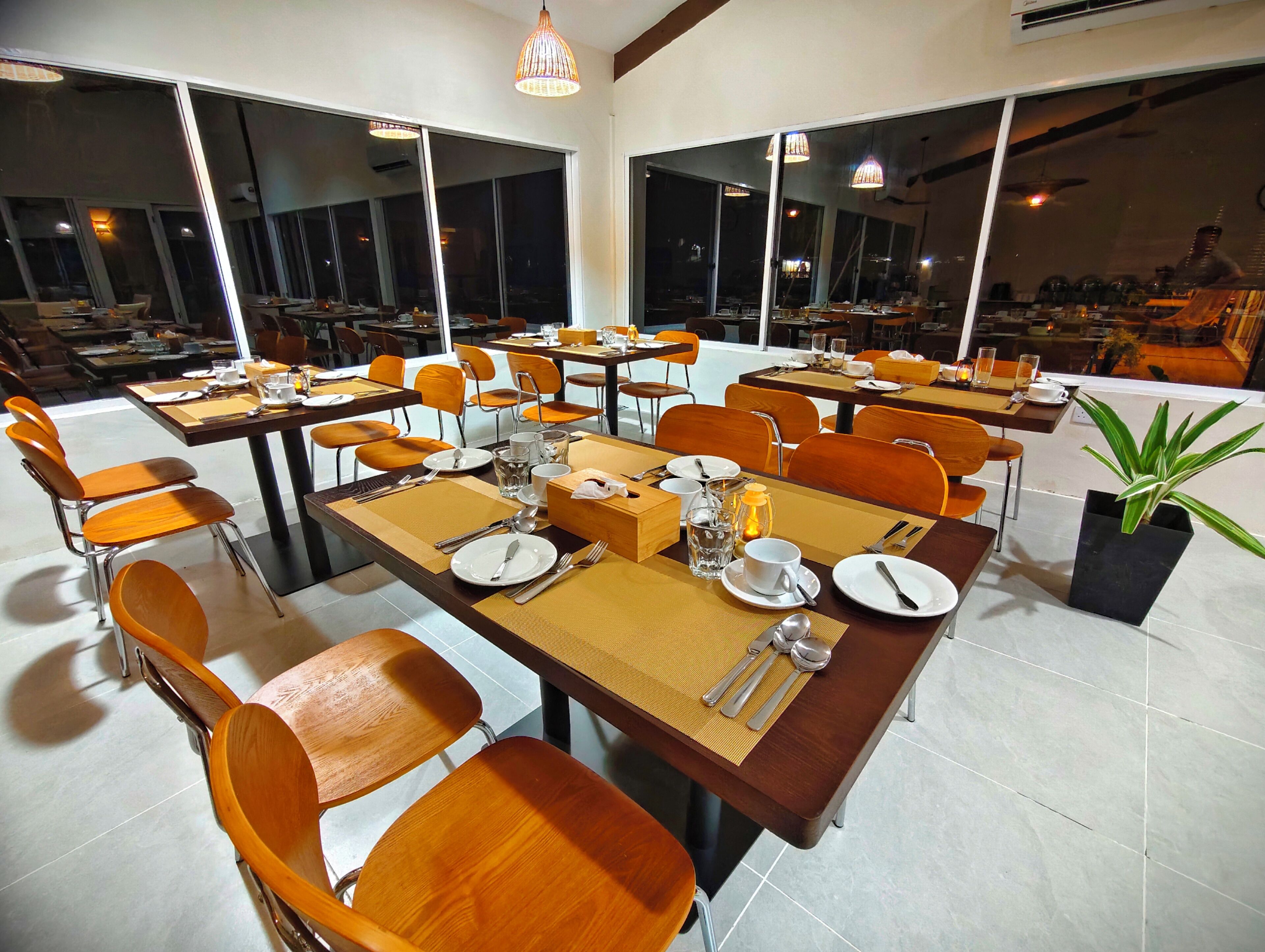 A spacious dining area with multiple tables and chairs, large windows, and a potted plant.