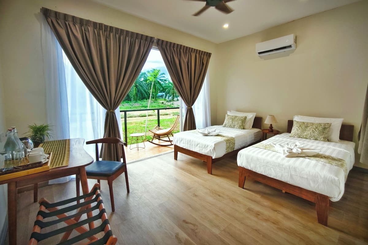 Hotelkamer met twee bedden, een plafondventilator, een airconditioningapparaat, een eettafel met stoelen en uitzicht op groen buiten.