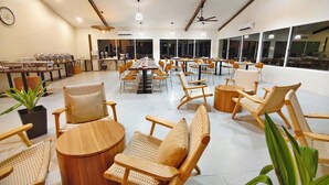 Un vaste espace dédié à la salle à manger, avec de grandes tables et des chaises en osier, de grandes fenêtres et des ventilateurs de plafond.