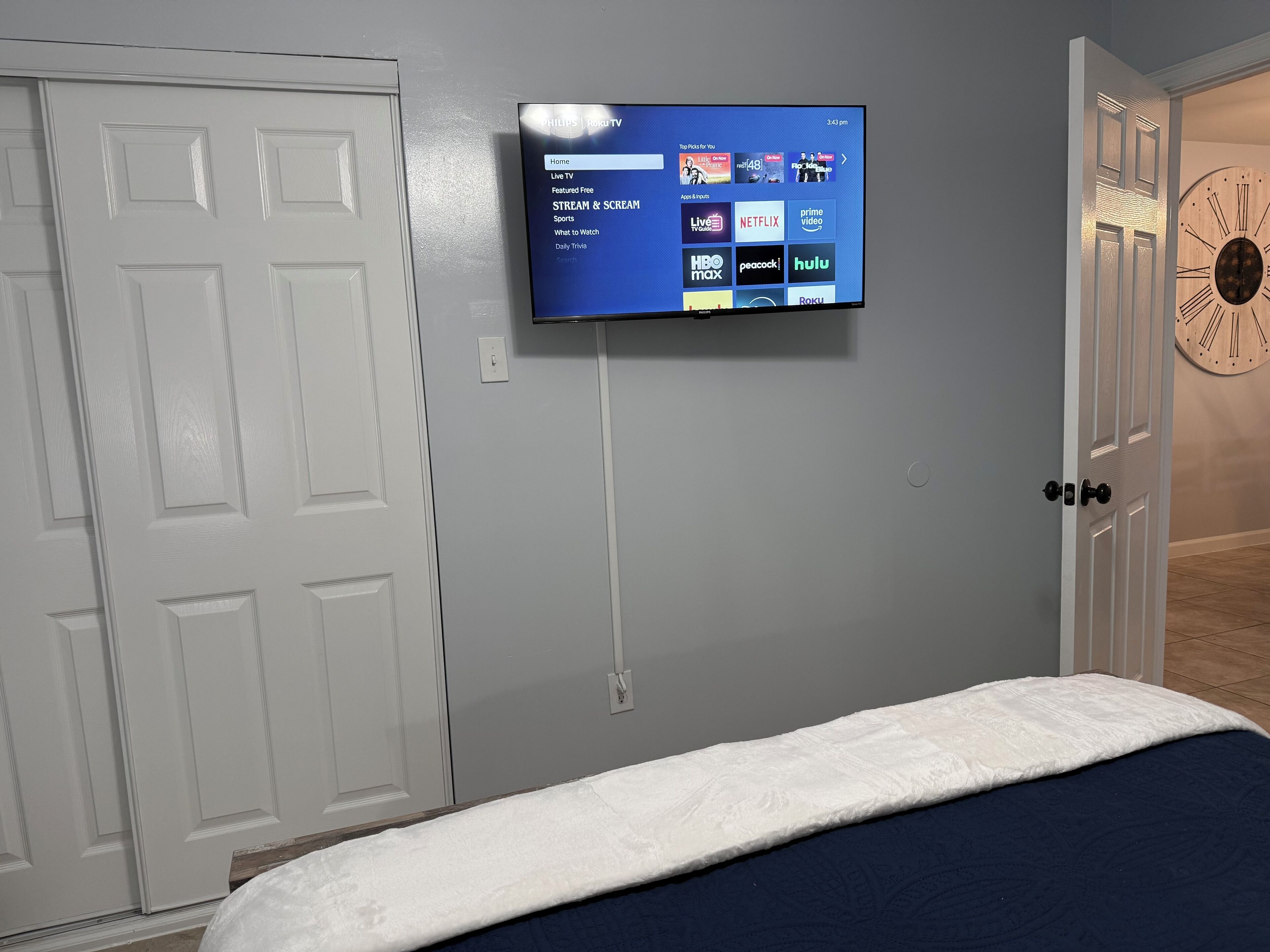Een slaapkamer met een bed, een wandtelevisie waarop een streamingdienst draait, een witte deur en een klok aan de muur.