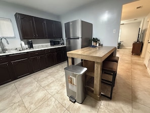 A kitchen with dark cabinets, a stainless steel refrigerator, a wooden table, and a trash bin.
