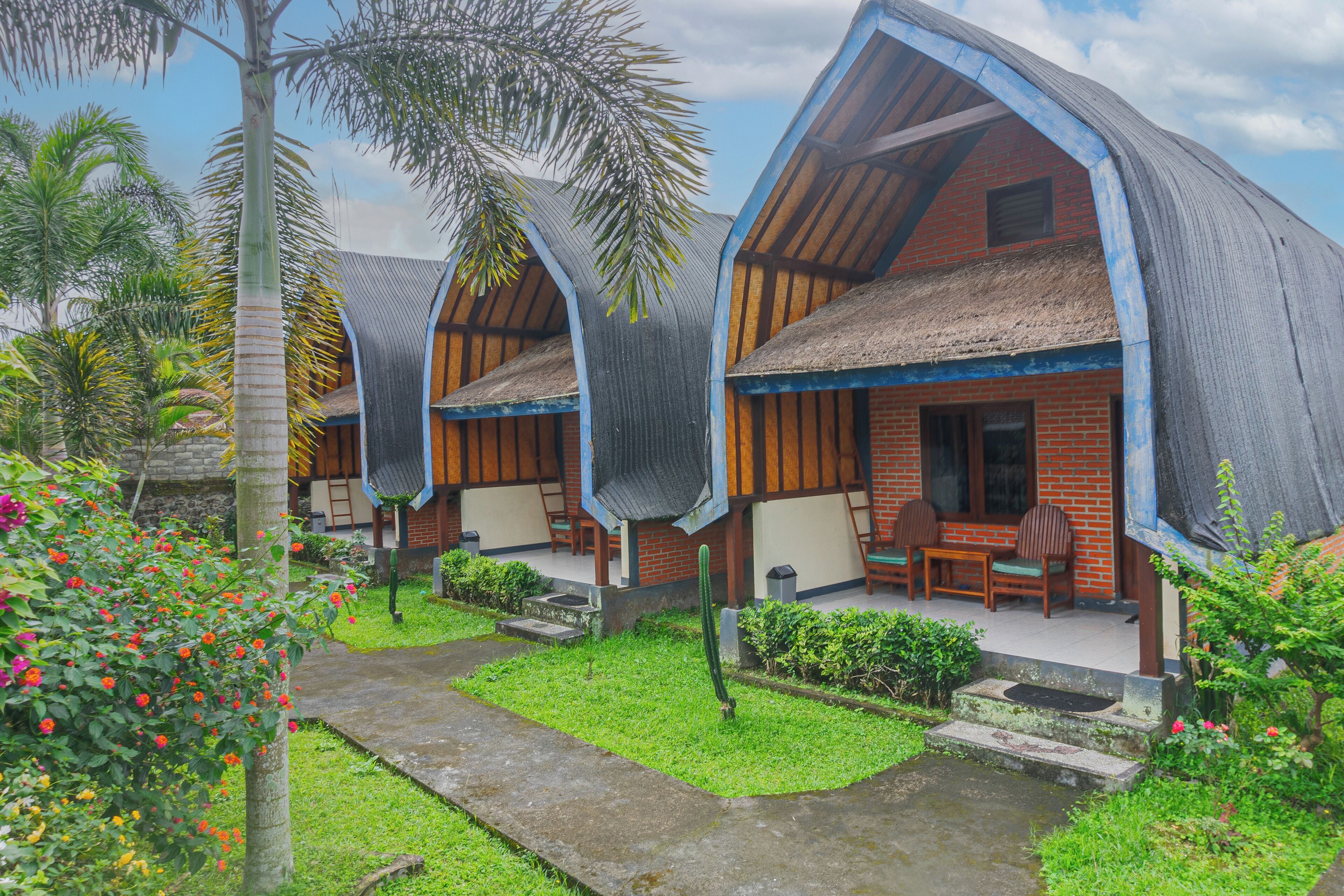 Un hotel de estilo bungalow con techos de paja y vigas de madera, que cuenta con un patio con mobiliario exterior y abundante vegetación.