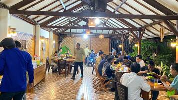 A restaurant with a tiled floor and wooden beams on the ceiling. Tables are arranged for dining, and people are seated.