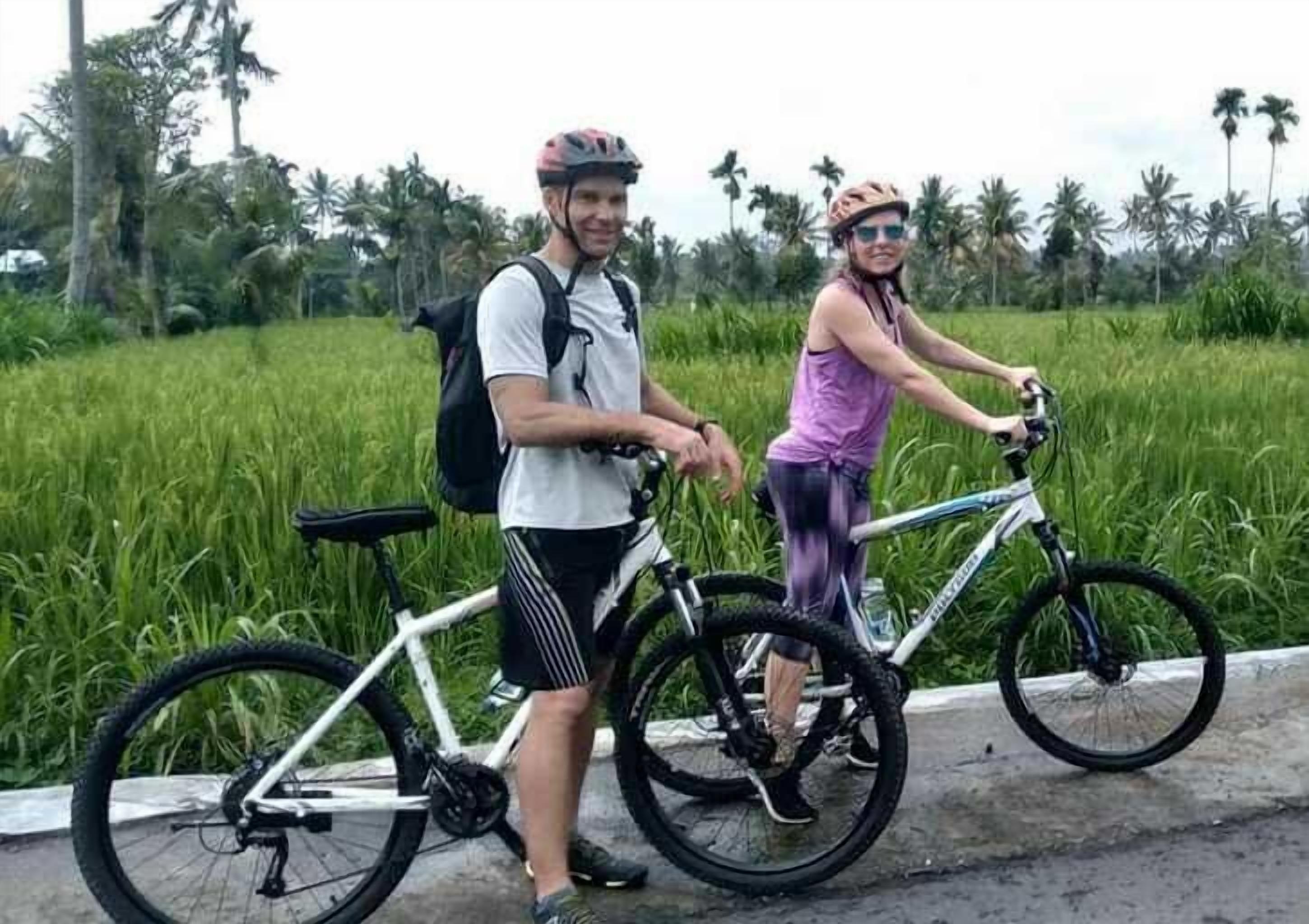Two people standing next to bicycles on a paved path with a green field and palm trees in the background.