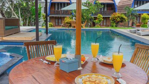 A poolside dining area with a wooden table, chairs, and a breakfast setting including plates, glasses of orange juice, and a basket. - Green Orry Inn Villas & Resort (Tetebatu)