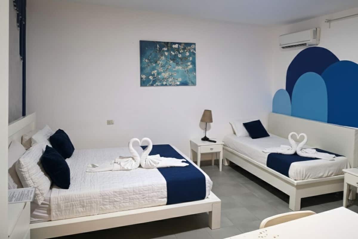 Ein Hotelzimmer mit zwei Betten, jeweils mit weißem Kopfteil, blauem Ziertekstoff und einer Anordnung aus Handtüchern in Form eines weißen Schwans. An der Wand hängt ein blaues Bild, und neben einem Bett steht ein kleiner Tisch mit einer Lampe.