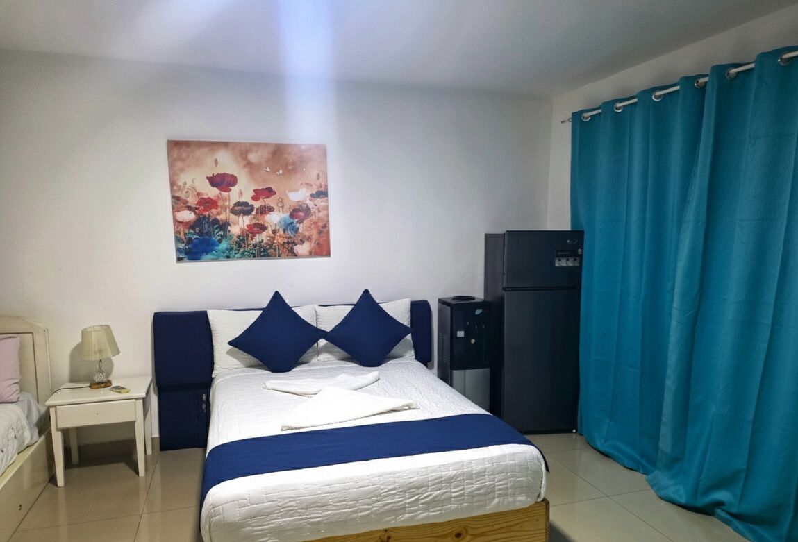 A hotel room with a bed, a nightstand, a lamp, a painting, a refrigerator, and a curtain.